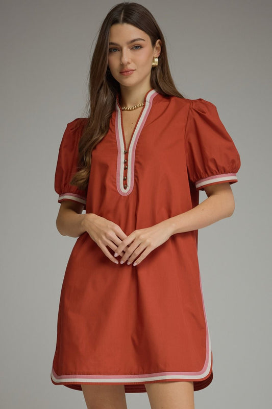 A woman modeling a terracotta-colored puff-sleeve mini dress with white contrast trim along the neckline, sleeve cuffs, and hem, set against a solid gray studio background.
