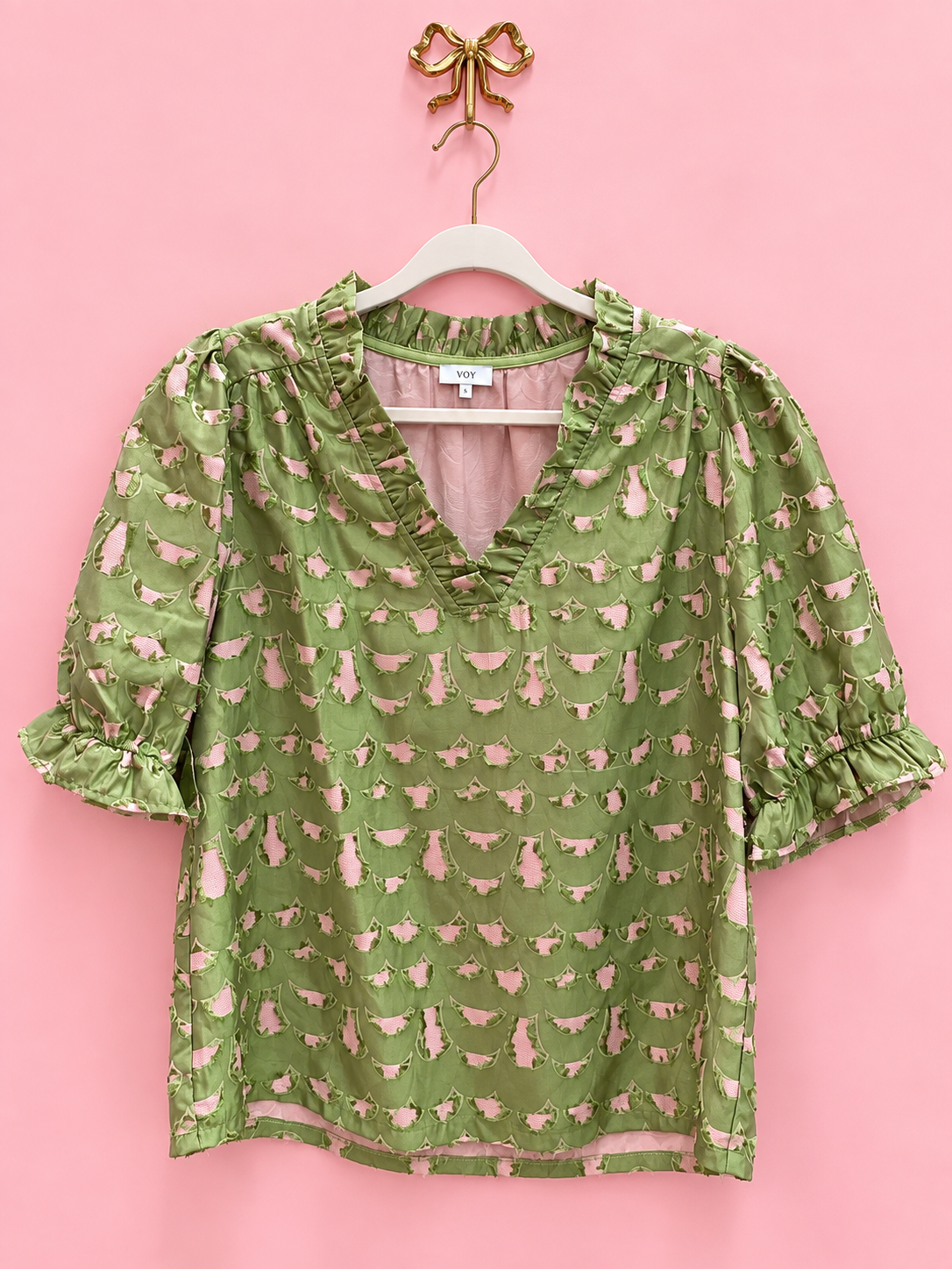 An olive green short-sleeve blouse with a pink sheep print, featuring ruffled trim at the V-neck and cuffs, hanging on a white hanger against a solid pink background.