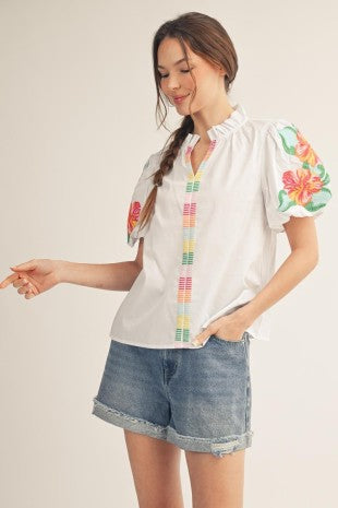 Woman wearing a white blouse with colorful floral sleeves and denim shorts on a plain background