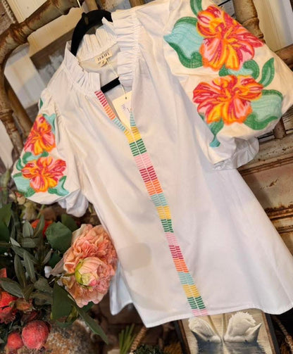 White blouse with colorful floral embroidery on a wooden surface with flowers.