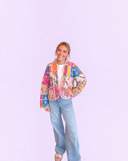 A woman modeling a colorful patchwork quilted jacket with a white top and light blue jeans, standing against a solid light purple background.