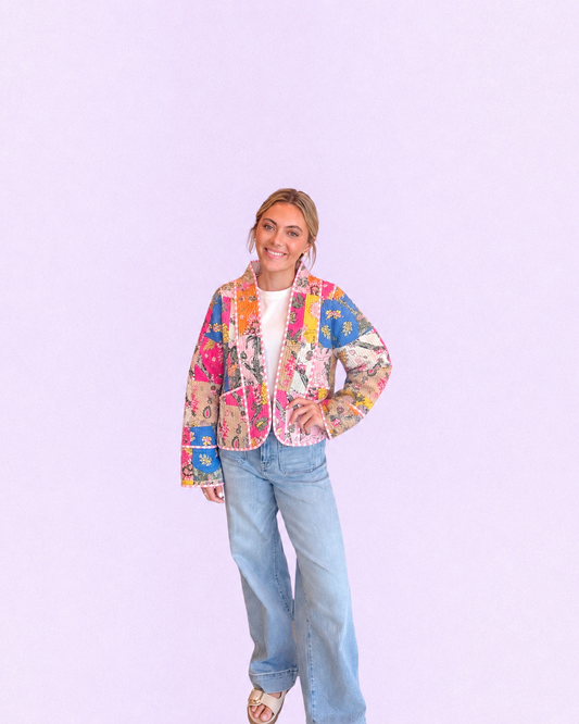 A woman modeling a colorful patchwork quilted jacket with a white top and light blue jeans, standing against a solid light purple background.