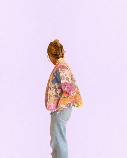 A person shown from the back wearing a colorful patchwork print jacket and light blue jeans against a solid light purple background.