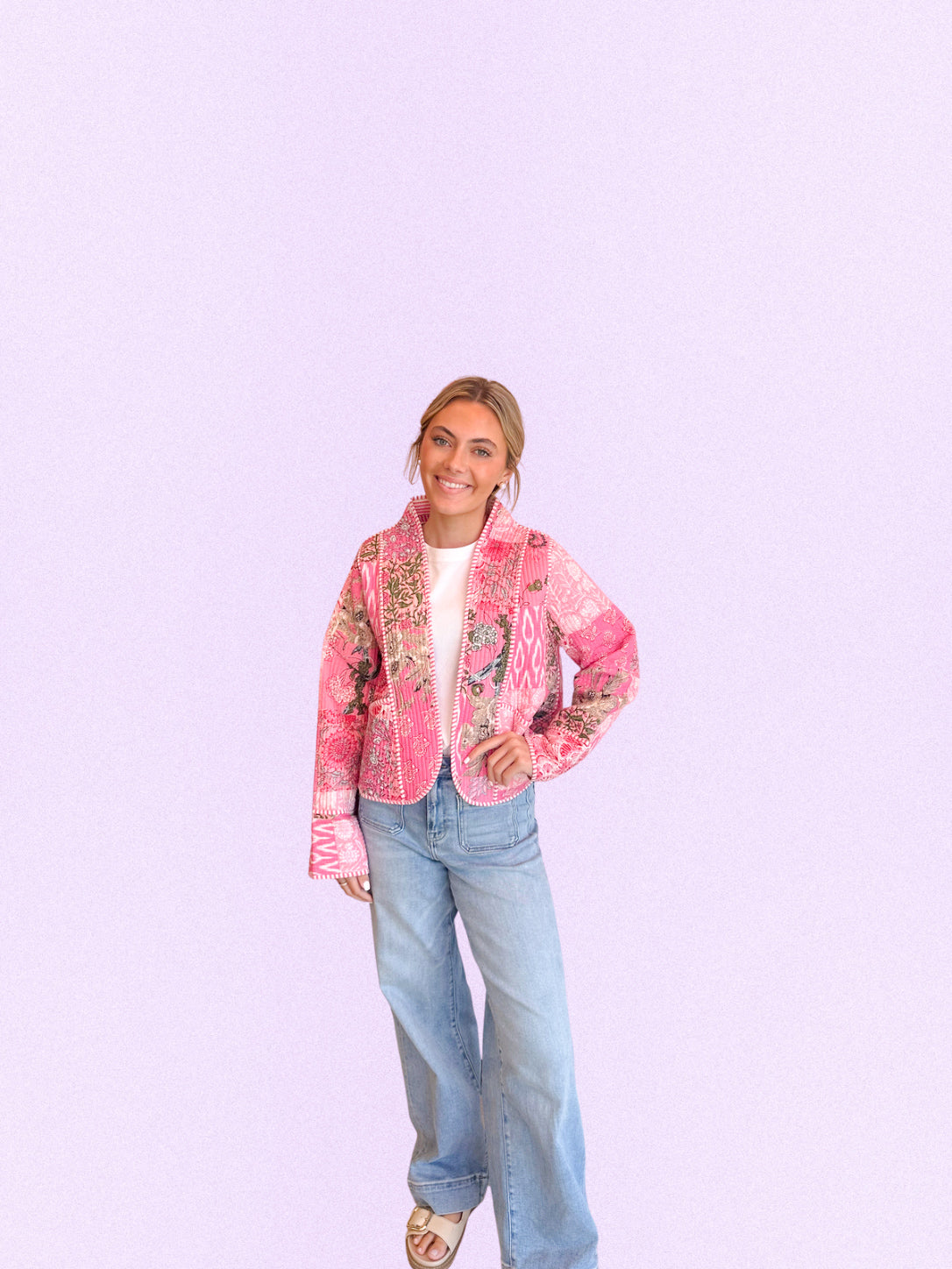 A woman modeling a pink embroidered jacket with floral and bird patterns, paired with light blue jeans and a white top, standing against a solid light purple background.