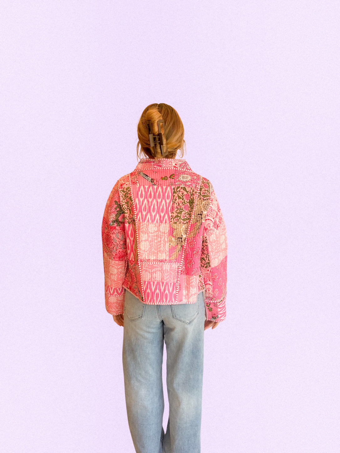 A person viewed from behind wearing a pink patchwork print jacket and light blue jeans against a solid pale lavender background.