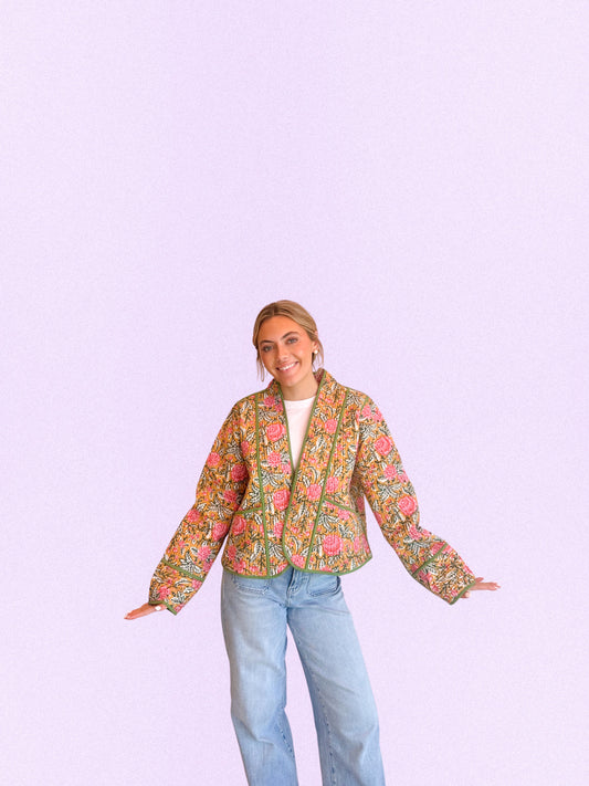 A woman modeling a cropped, long-sleeve floral jacket with a vibrant pink, green, and orange pattern, paired with light blue jeans, set against a solid light purple background.