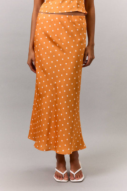 An orange midi skirt with a white polka dot pattern, shown on a model wearing a matching orange top and white flip-flop sandals against a neutral gray background.