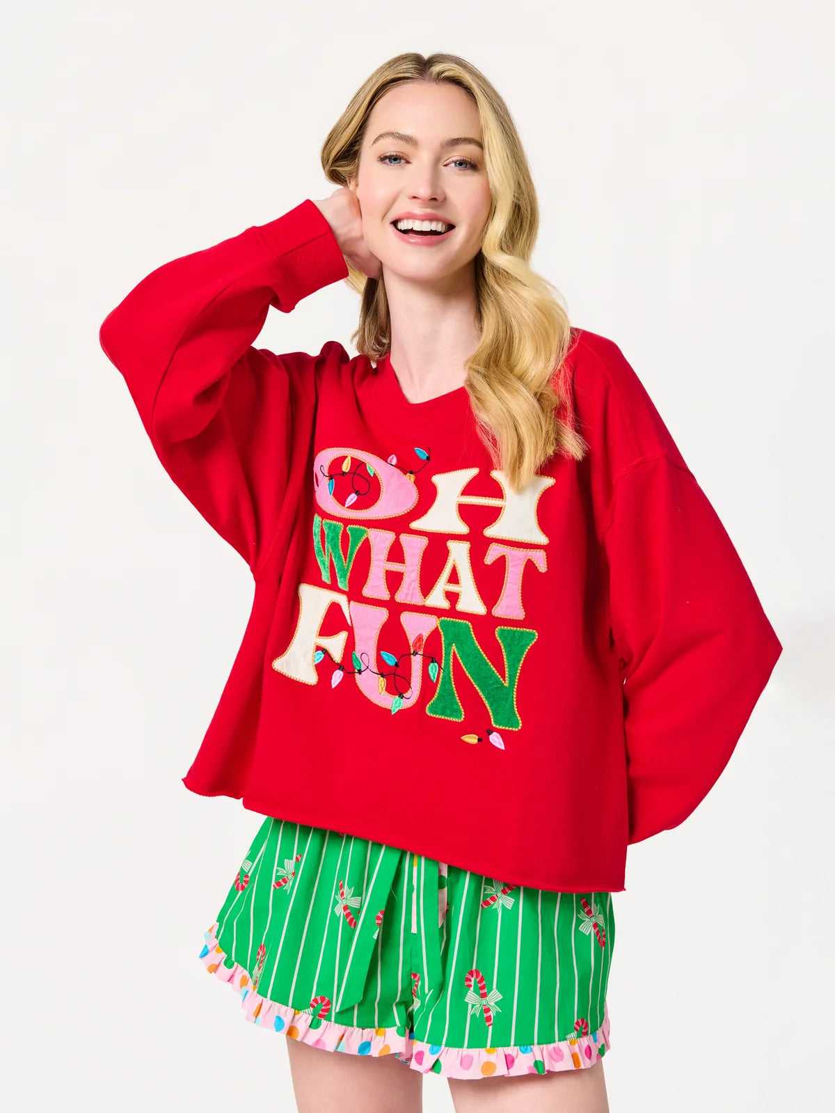 A woman modeling a red oversized hoodie with colorful 'OH WHAT FUN' text on the front, paired with green striped shorts with a candy cane print, against a plain white background.
