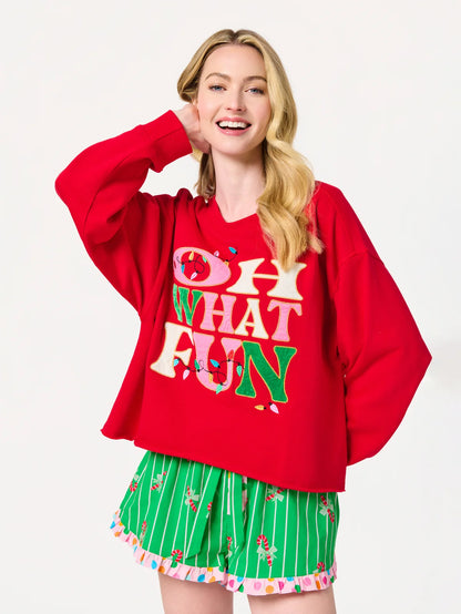 A woman modeling a red oversized hoodie with colorful 'OH WHAT FUN' text on the front, paired with green striped shorts with a candy cane print, against a plain white background.