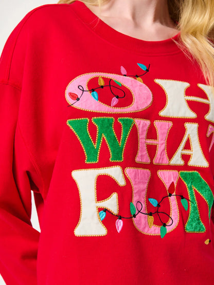 A close-up of a bright red crewneck sweatshirt featuring a large, colorful embroidered graphic that reads 'OH WHAT FUN' in pink, green, and cream letters, decorated with light bulb outlines.