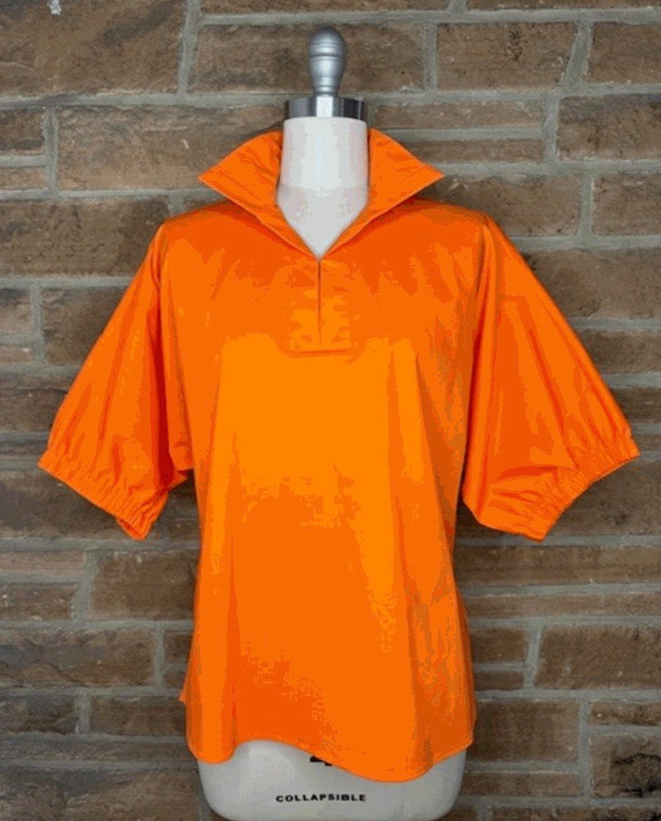 Bright orange short-sleeve collared pullover top displayed on a white mannequin torso against a brown brick wall background.