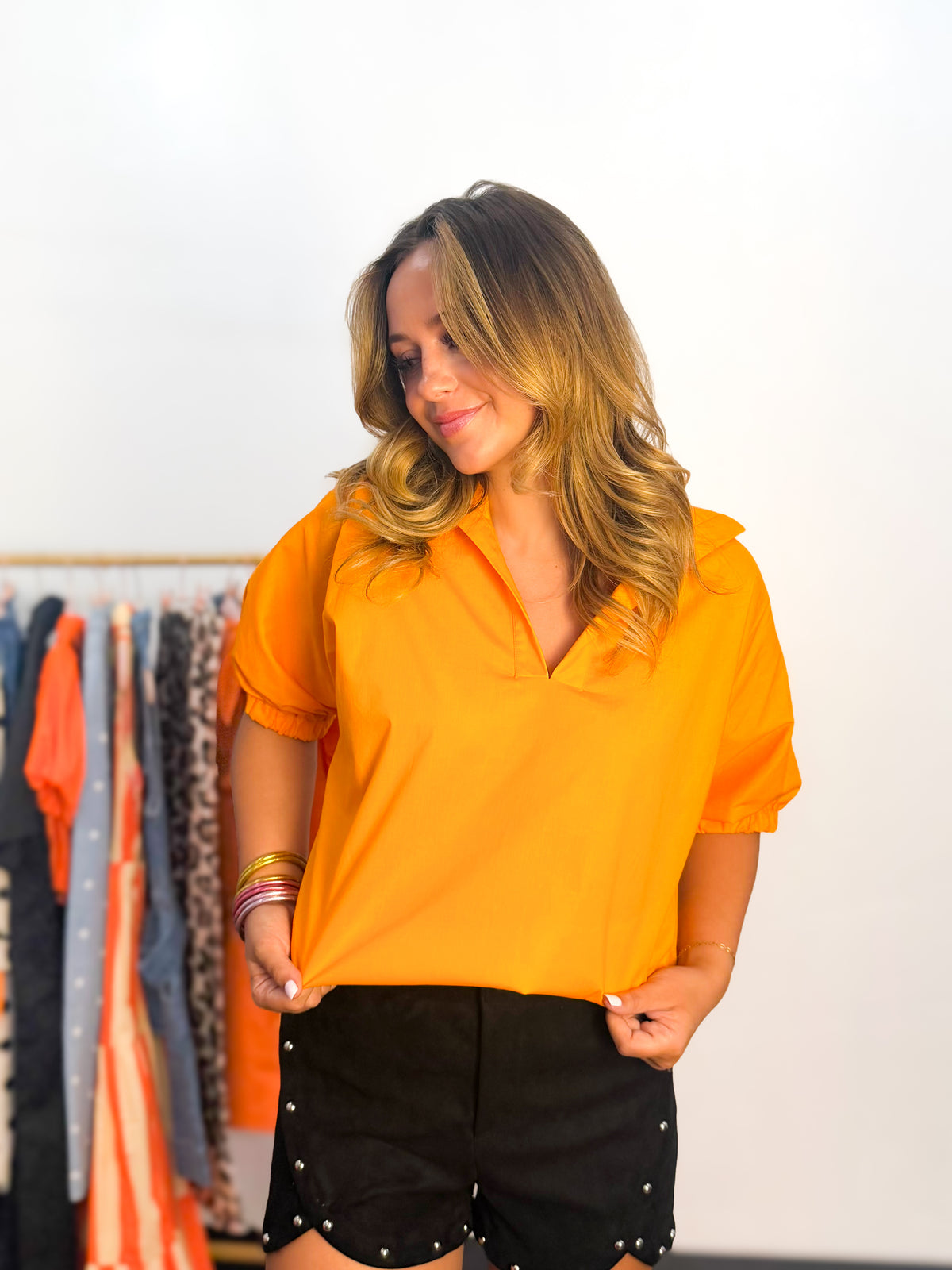 A woman wearing a bright orange short-sleeve collared blouse and black shorts with silver studs, standing in front of a white wall with a clothing rack visible in the background.