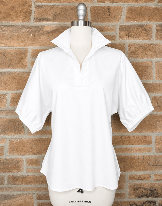 A white short-sleeve collared blouse with a partial zip and elasticized cuffs, displayed on a mannequin torso against a brown brick wall background.