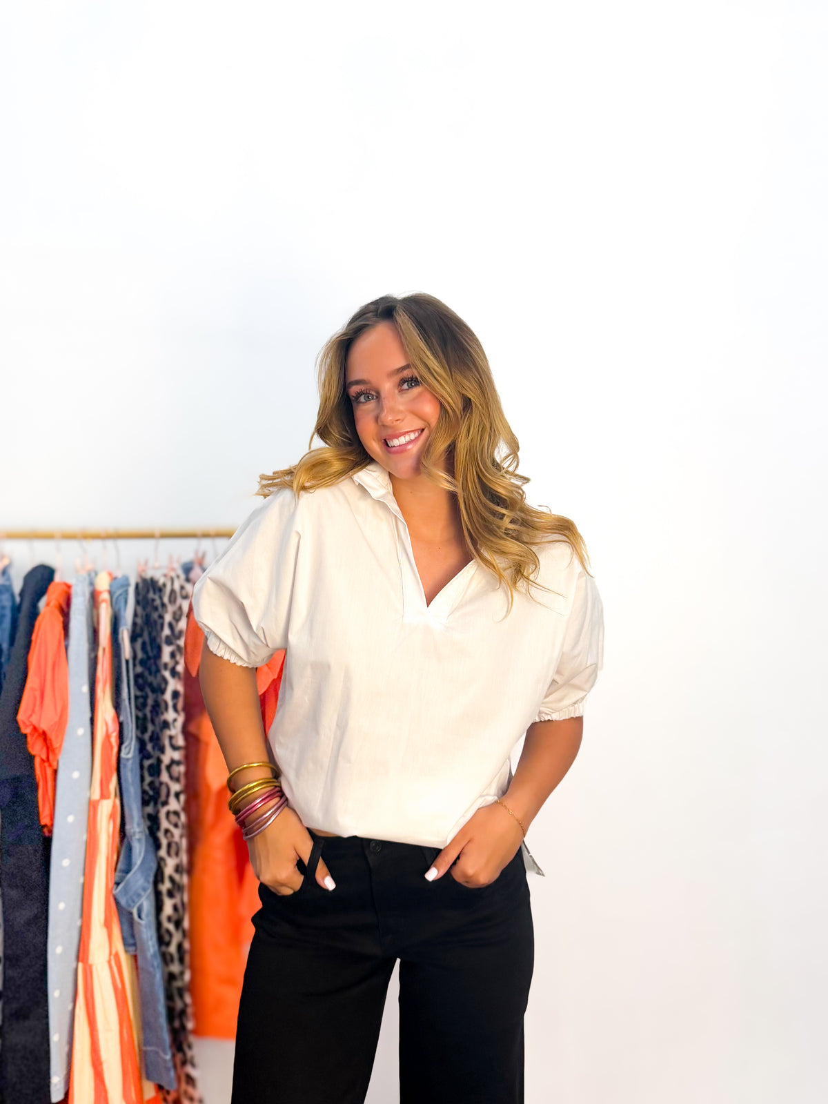 A woman smiling and posing in a white short-sleeve button-up blouse with black pants, standing in front of a white wall with a clothing rack visible on the left.