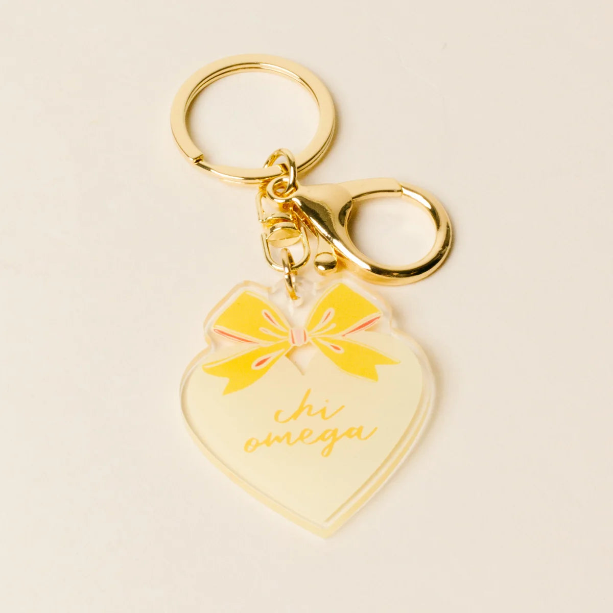 A gold-tone metal keychain with a heart-shaped acrylic charm. The charm features a yellow bow graphic and the cursive text 'chi omega' in gold, set against a clean, light beige background.
