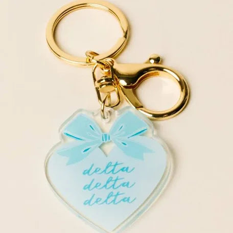 A heart-shaped acrylic keychain charm in light blue with a matching blue bow graphic and the word 'delta' written three times in script, attached to gold-tone metal rings and clasp on a light beige background.