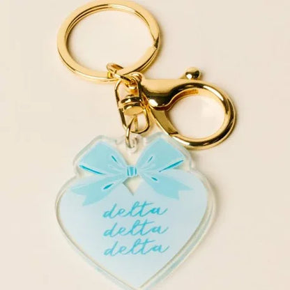 A heart-shaped acrylic keychain charm in light blue with a matching blue bow graphic and the word 'delta' written three times in script, attached to gold-tone metal rings and clasp on a light beige background.