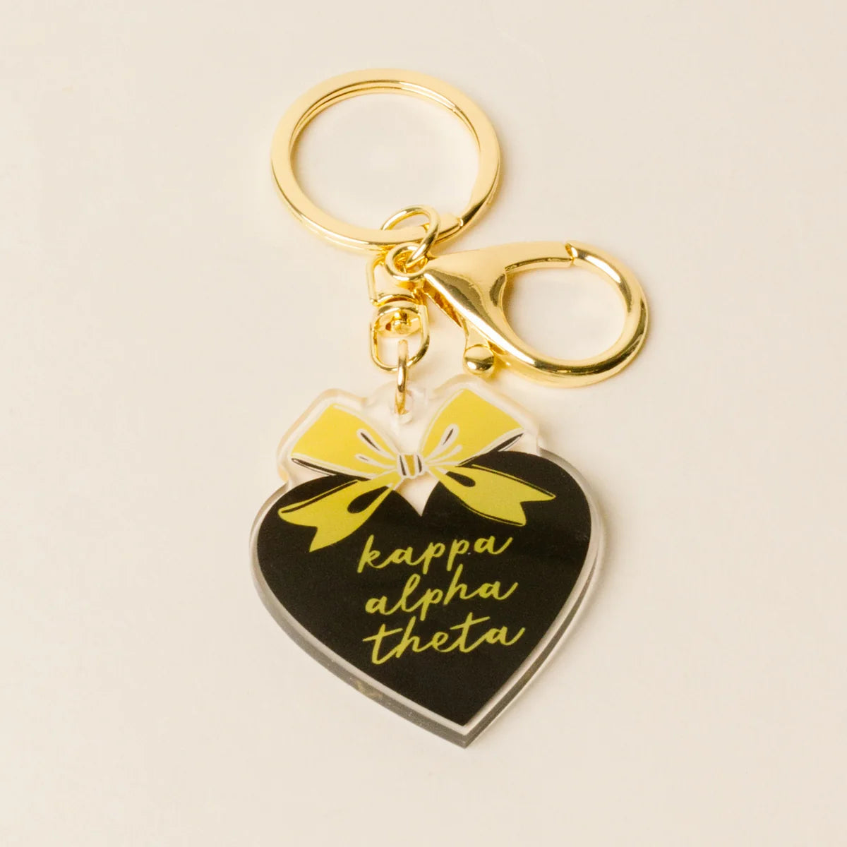 A heart-shaped acrylic keychain charm for Kappa Alpha Theta, featuring a black face with gold lettering, a yellow bow graphic, and a gold metal key ring and clasp on a light beige background.