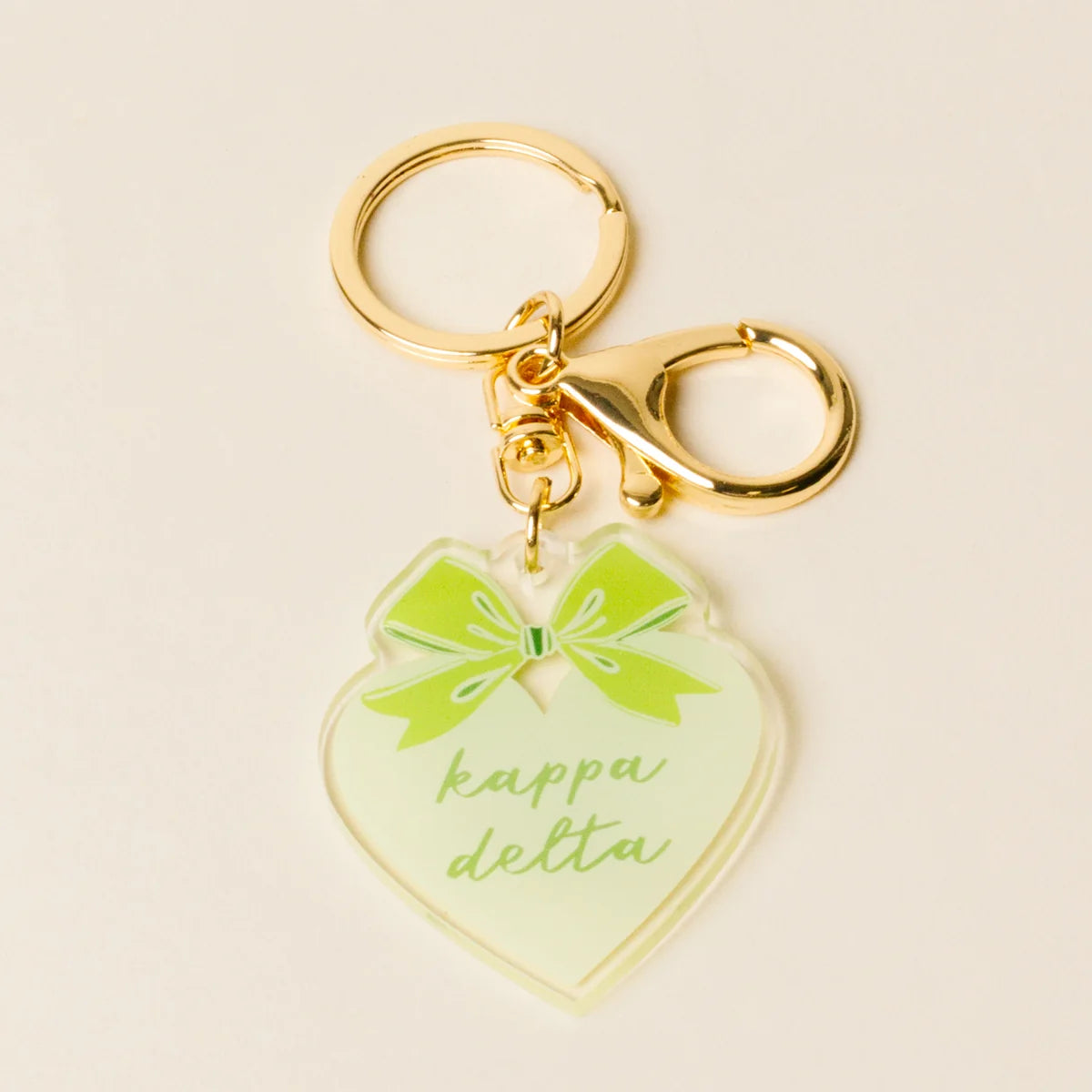 A heart-shaped acrylic keychain charm in pale green with a bright green bow graphic and the words 'kappa delta' in green script, attached to gold-tone metal hardware on a light beige background.