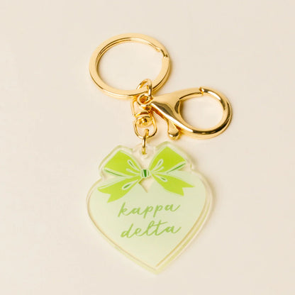 A heart-shaped acrylic keychain charm in pale green with a bright green bow graphic and the words 'kappa delta' in green script, attached to gold-tone metal hardware on a light beige background.