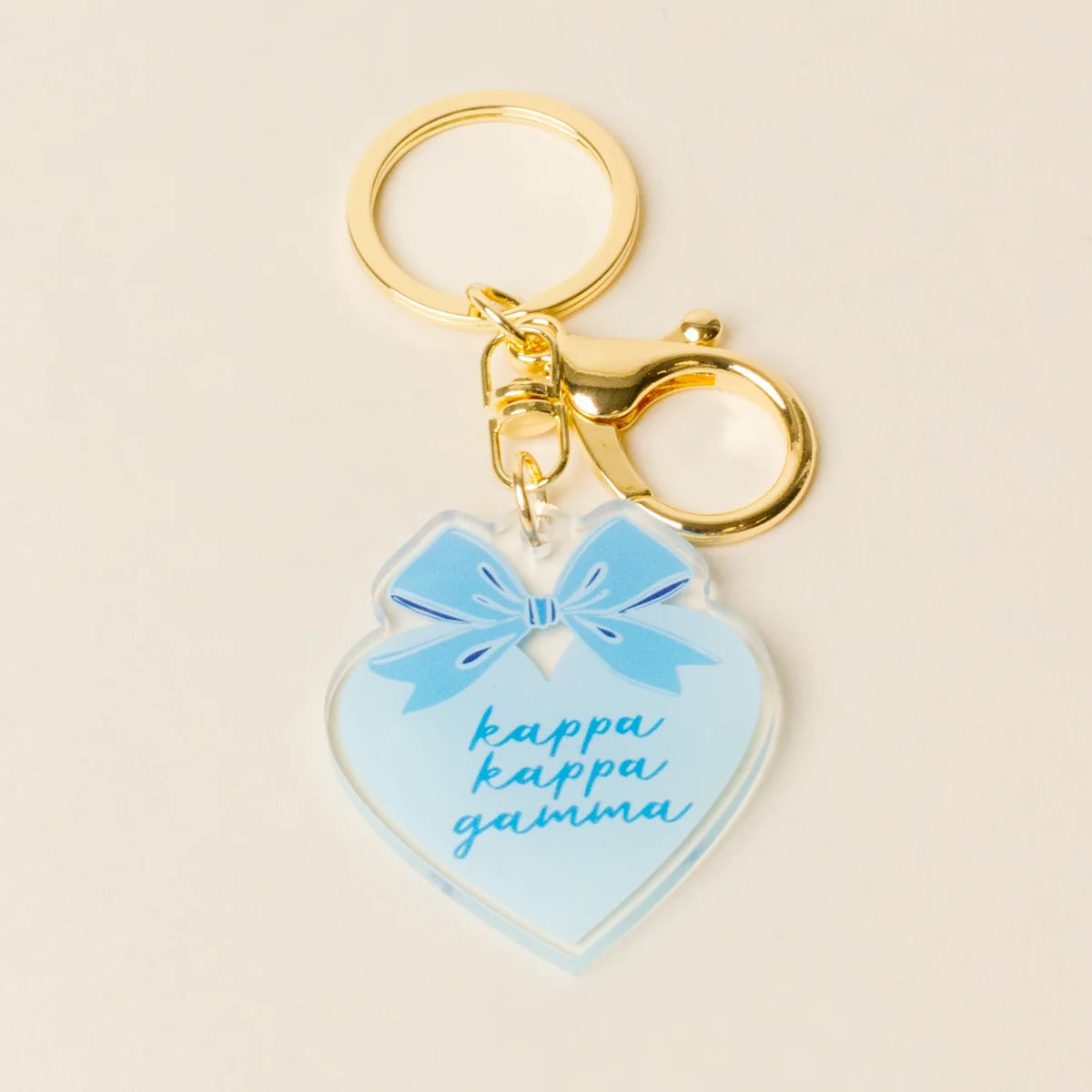 A heart-shaped acrylic keychain with a light blue bow graphic and the words 'kappa kappa gamma' in blue script, attached to gold metal rings on a light beige background.