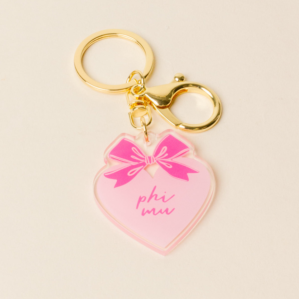 A heart-shaped acrylic keychain charm in light pink with a bright pink bow graphic and the words 'phi mu' in pink script, attached to gold-tone metal hardware on a light beige background.