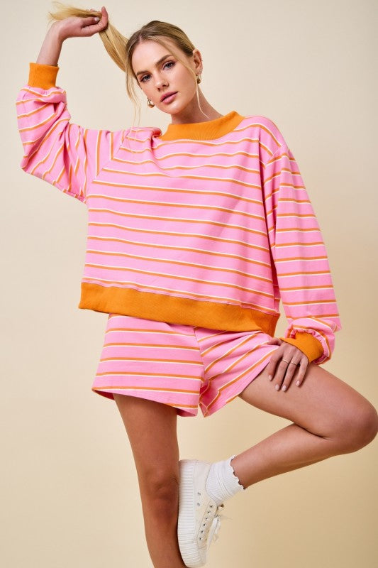 A woman modeling a pink and orange striped long-sleeve sweatshirt and matching shorts set against a solid beige background.