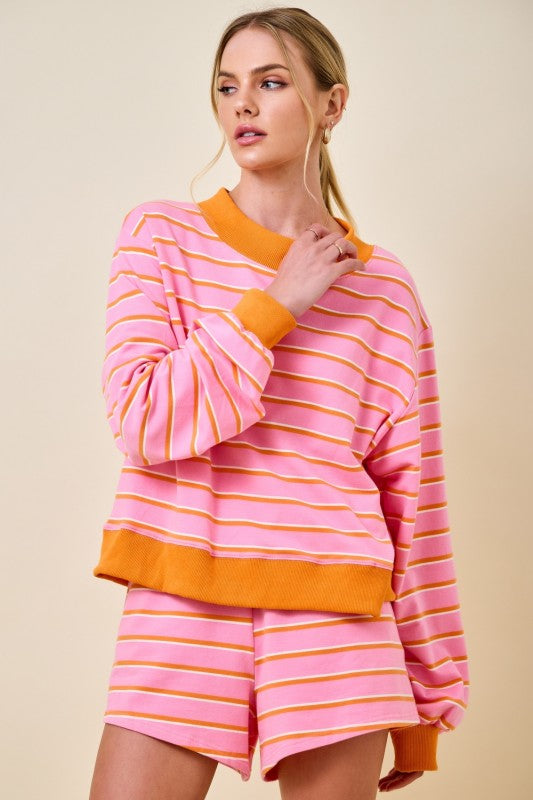 A woman modeling a pink and orange striped lounge set consisting of a long-sleeve sweatshirt and matching shorts, against a solid beige background.