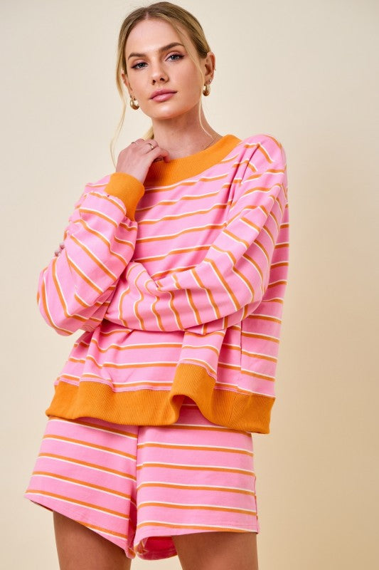 A woman modeling a pink and orange striped lounge set consisting of a long-sleeve top and matching shorts, set against a solid beige background.