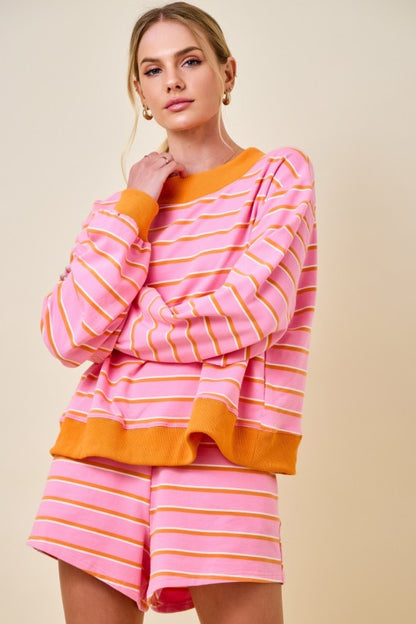 A woman modeling a pink and orange striped lounge set consisting of a long-sleeve top and matching shorts, set against a solid beige background.