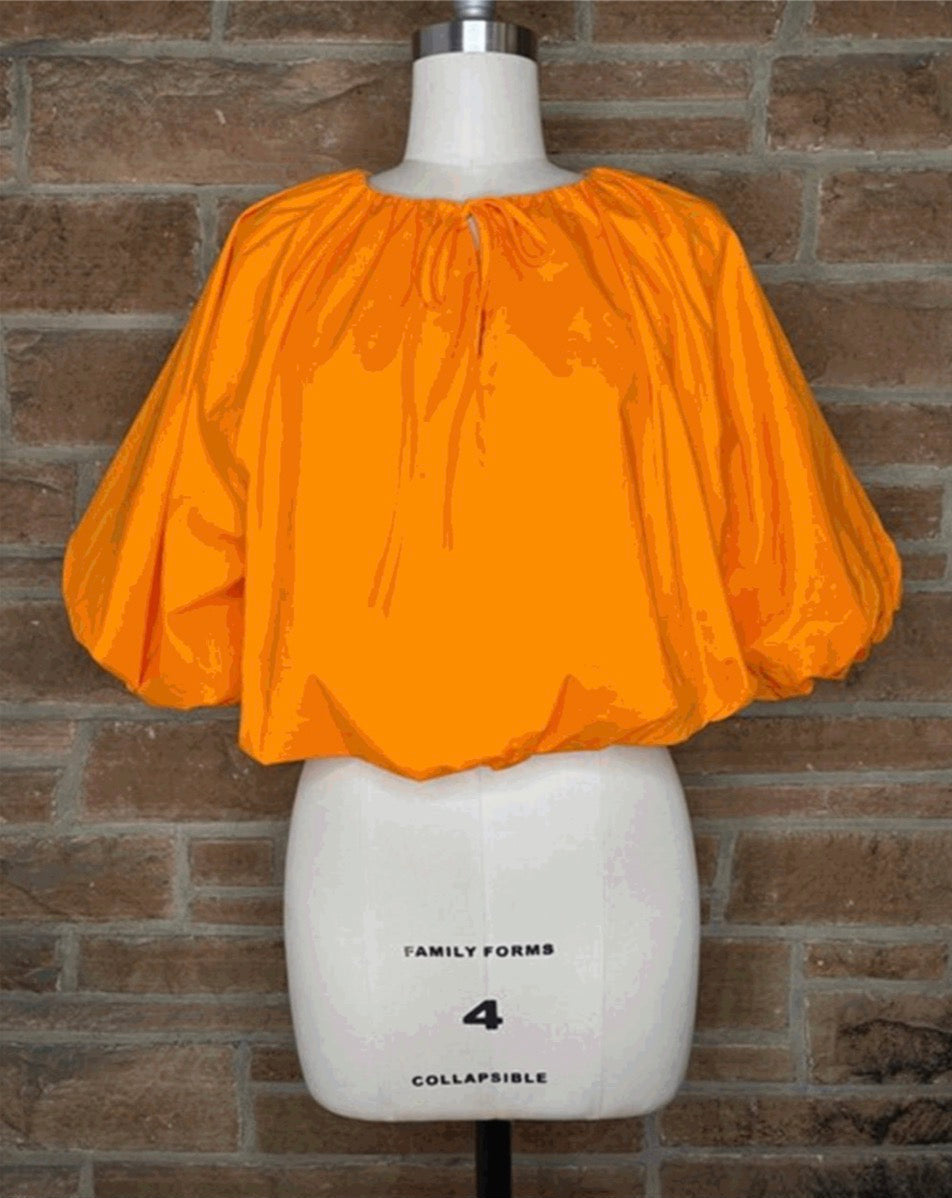 Bright orange short puff-sleeve top displayed on a white dress form mannequin against a brown brick wall background.