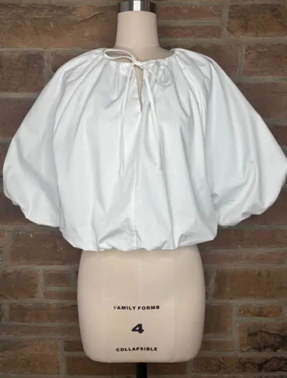 A white puff-sleeve blouse with a tie-neck detail, displayed on a beige mannequin torso against a brown brick wall background.