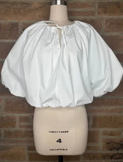 A white puff-sleeve blouse with a tie-neck detail, displayed on a beige mannequin torso against a brown brick wall background.