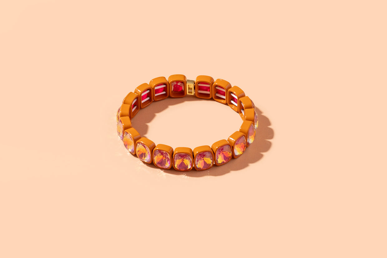 A gold-toned stretch bracelet featuring alternating rectangular pinkish-red stones and smaller round iridescent pink and orange stones, shown on a solid peach background.