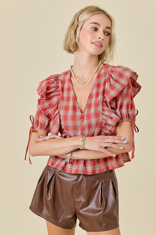 A woman modeling a red and white plaid short-sleeve blouse with ruffled shoulders and tie details, paired with brown faux-leather shorts, against a solid beige background.