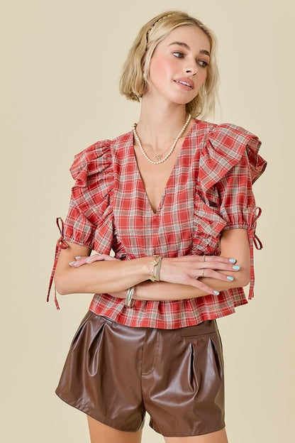 A woman modeling a red and white plaid short-sleeve blouse with ruffled shoulders and tie details, paired with brown faux-leather shorts, against a solid beige background.