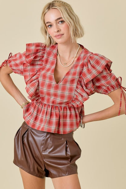 A woman modeling a red and white plaid short-sleeve blouse with ruffled shoulders and side ties, paired with brown faux-leather shorts, against a solid beige background.