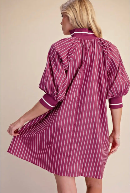 A woman shown from the back wearing a short puff-sleeve shirt dress with vertical magenta and white stripes, set against a solid beige background.
