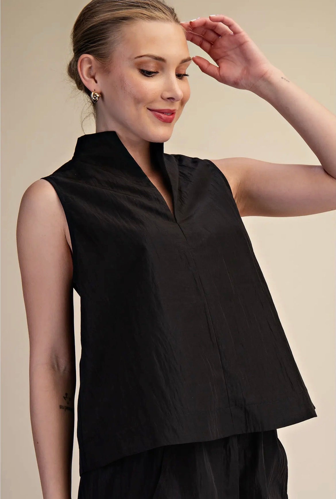 A woman wearing a sleeveless black high-neck top with a V-neckline, shown against a plain beige studio background.