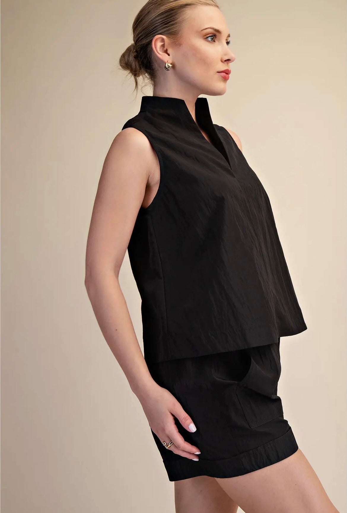 A woman modeling a sleeveless black high-neck top and matching black shorts against a plain beige studio background.