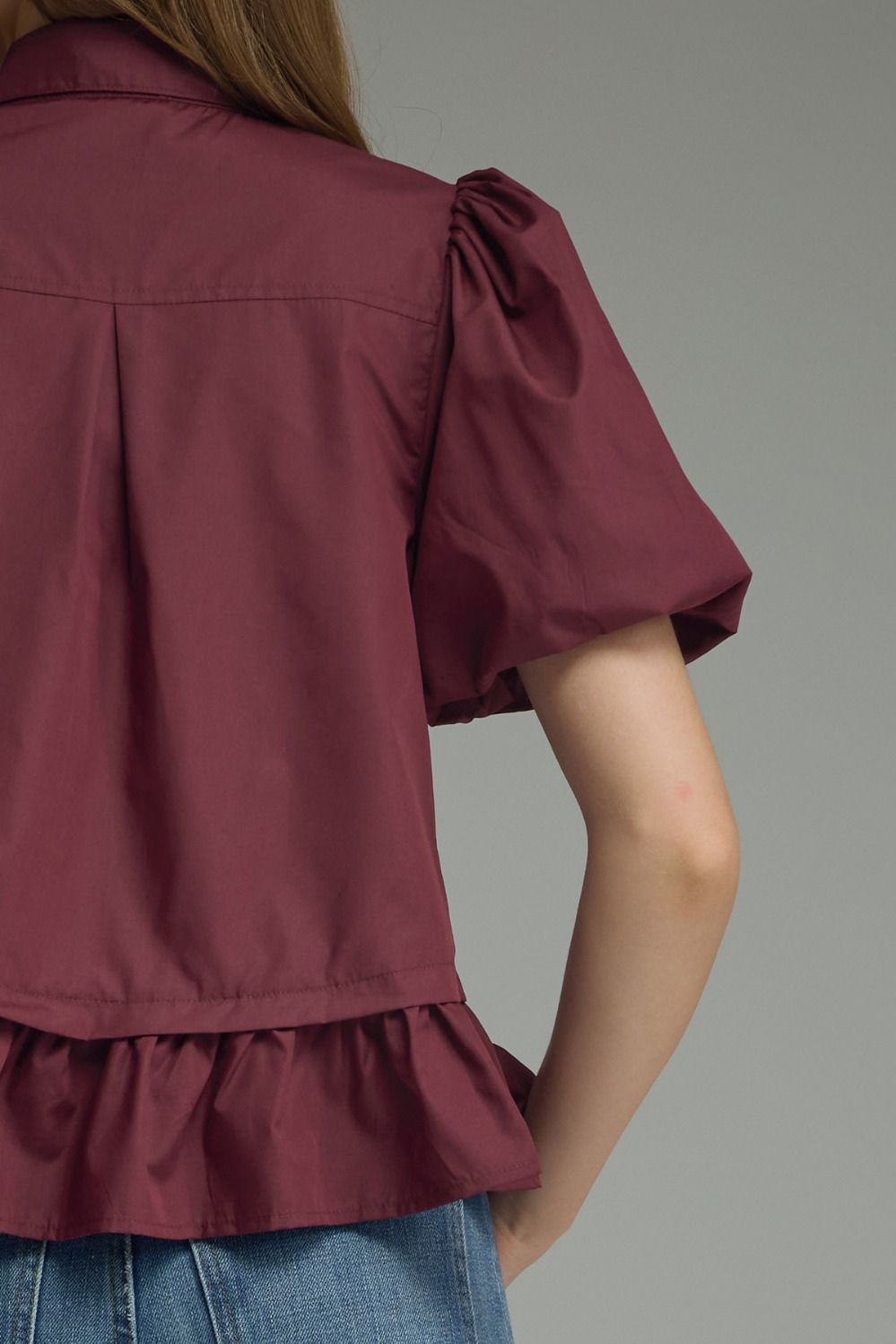 A close-up back view of a person wearing a short-sleeve burgundy blouse with puff sleeves and a ruffled hem, paired with blue jeans against a plain gray background.