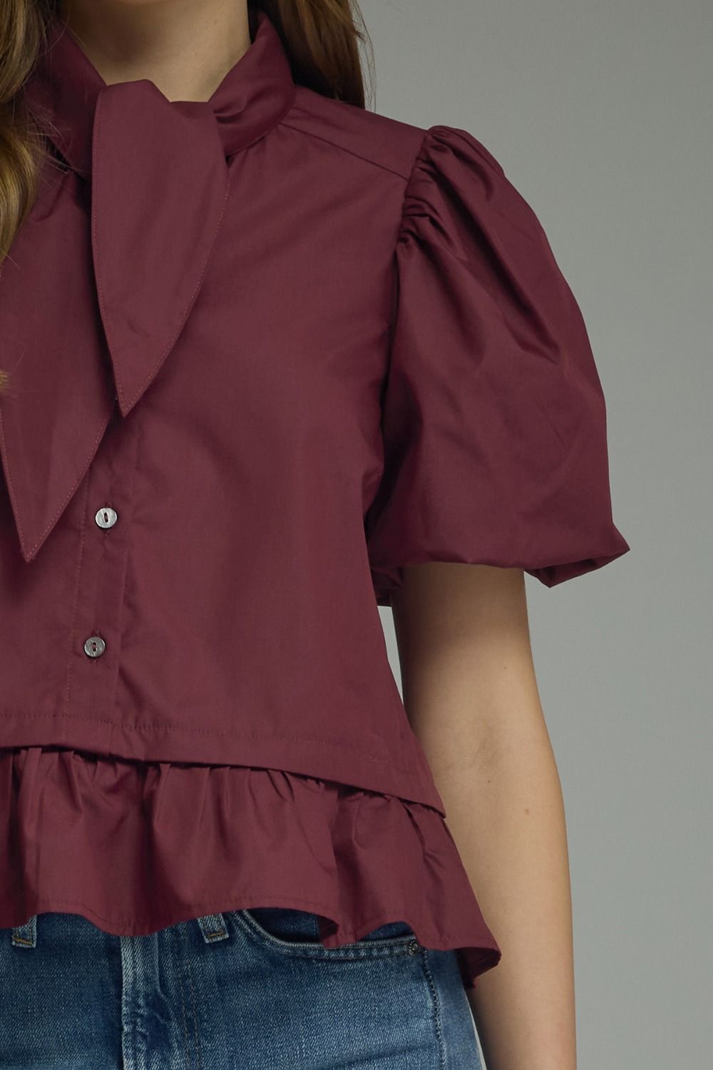 A close-up of a woman wearing a short-sleeve burgundy blouse with a large bow collar, puff sleeves, and a ruffled hem, paired with blue jeans against a plain gray background.