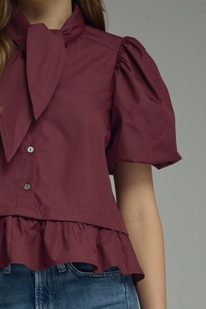 A close-up of a woman wearing a short-sleeve burgundy blouse with a large bow collar, puff sleeves, and a ruffled hem, paired with blue jeans against a plain gray background.