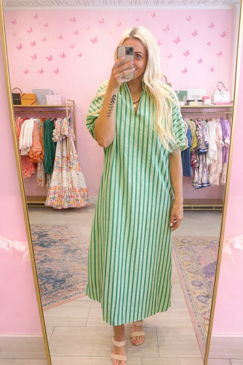 Woman in a green striped dress taking a mirror selfie in a store with pink walls and butterfly decorations.