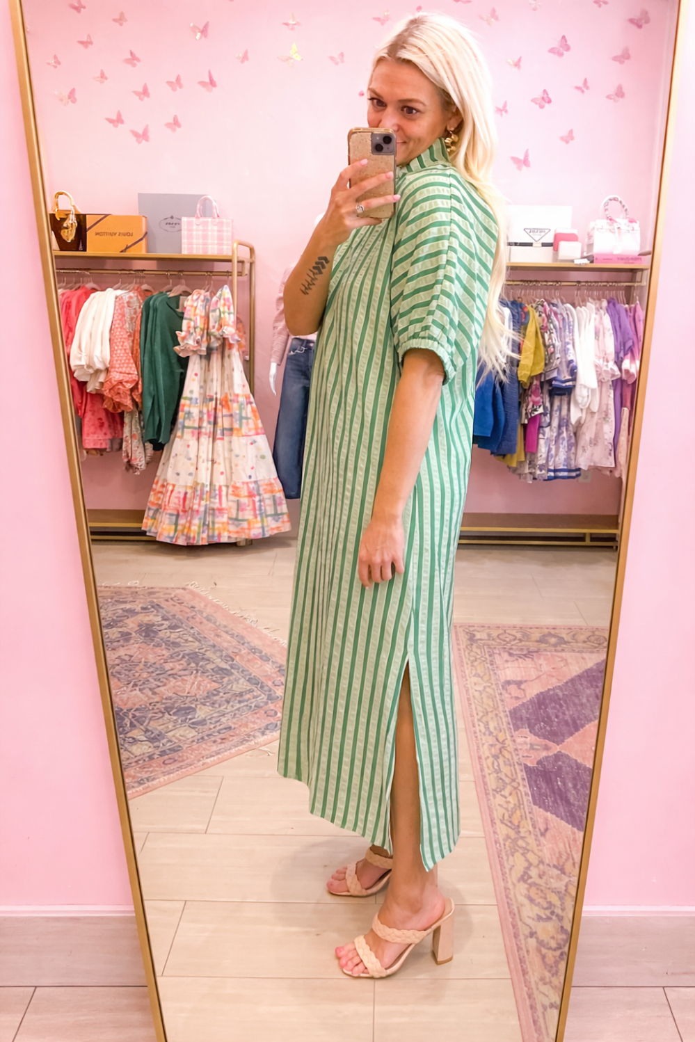 Woman in a green striped dress standing in front of a mirror in a pink room with clothing racks.