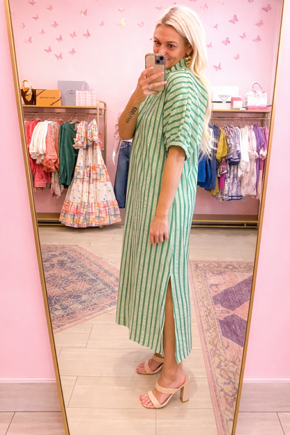 Woman in a green striped dress standing in front of a mirror in a pink room with clothing racks.