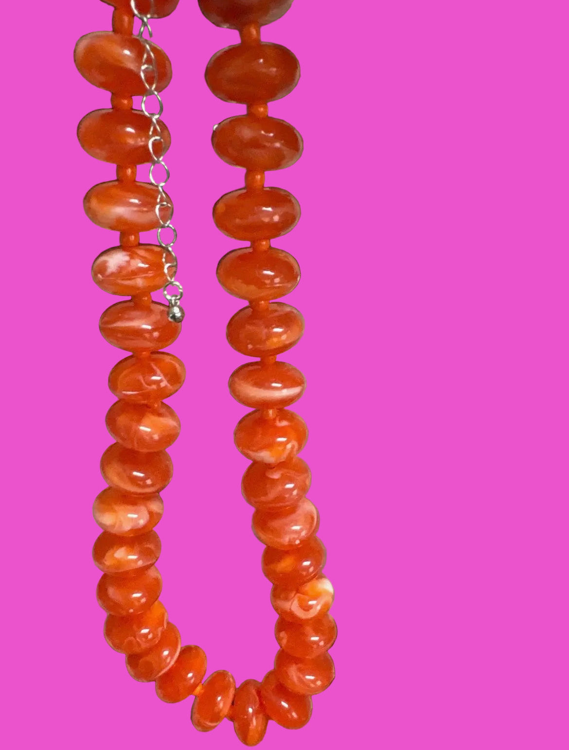 A person holding up a long strand of chunky orange beads, likely a necklace or bracelet, in an indoor setting. The beads are glossy and translucent, and a small silver clasp is visible at the top.