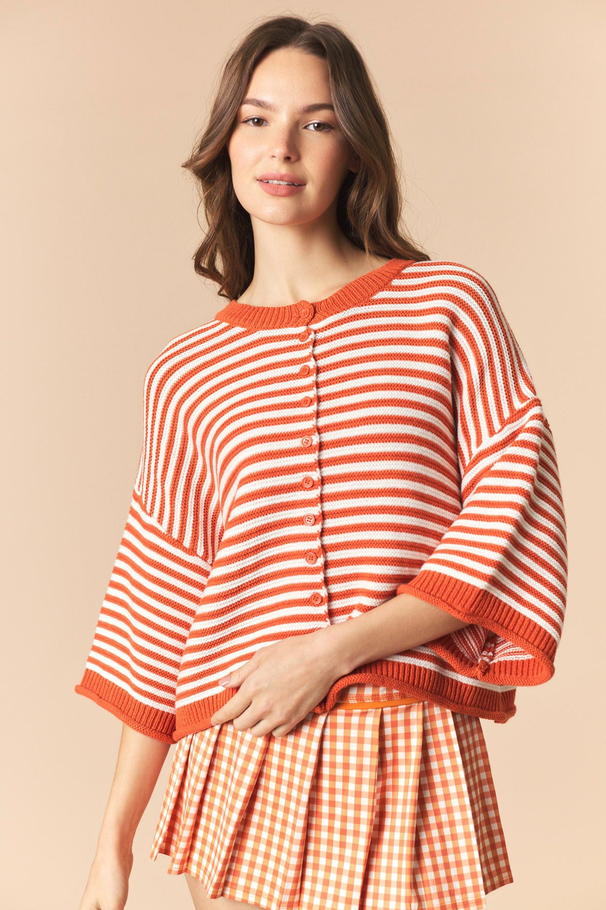 A woman wearing a short-sleeve knit top with orange and white horizontal stripes and a matching orange gingham pleated skirt, posed against a solid beige background.