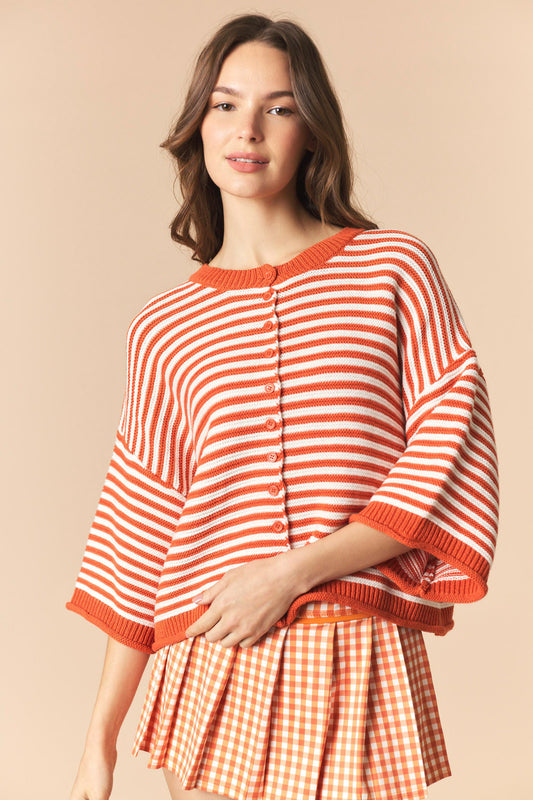 A woman wearing a short-sleeve knit top with orange and white horizontal stripes and a matching orange gingham pleated skirt, posed against a solid beige background.