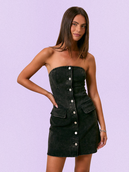 A woman modeling a strapless black denim mini dress with a button-down front and side pockets, set against a solid lavender background.